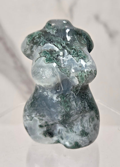 A small polished stone figurine shaped like a female torso, featuring dark green and white marbled patterning, displayed on a clear acrylic stand against a light marble-patterned background.