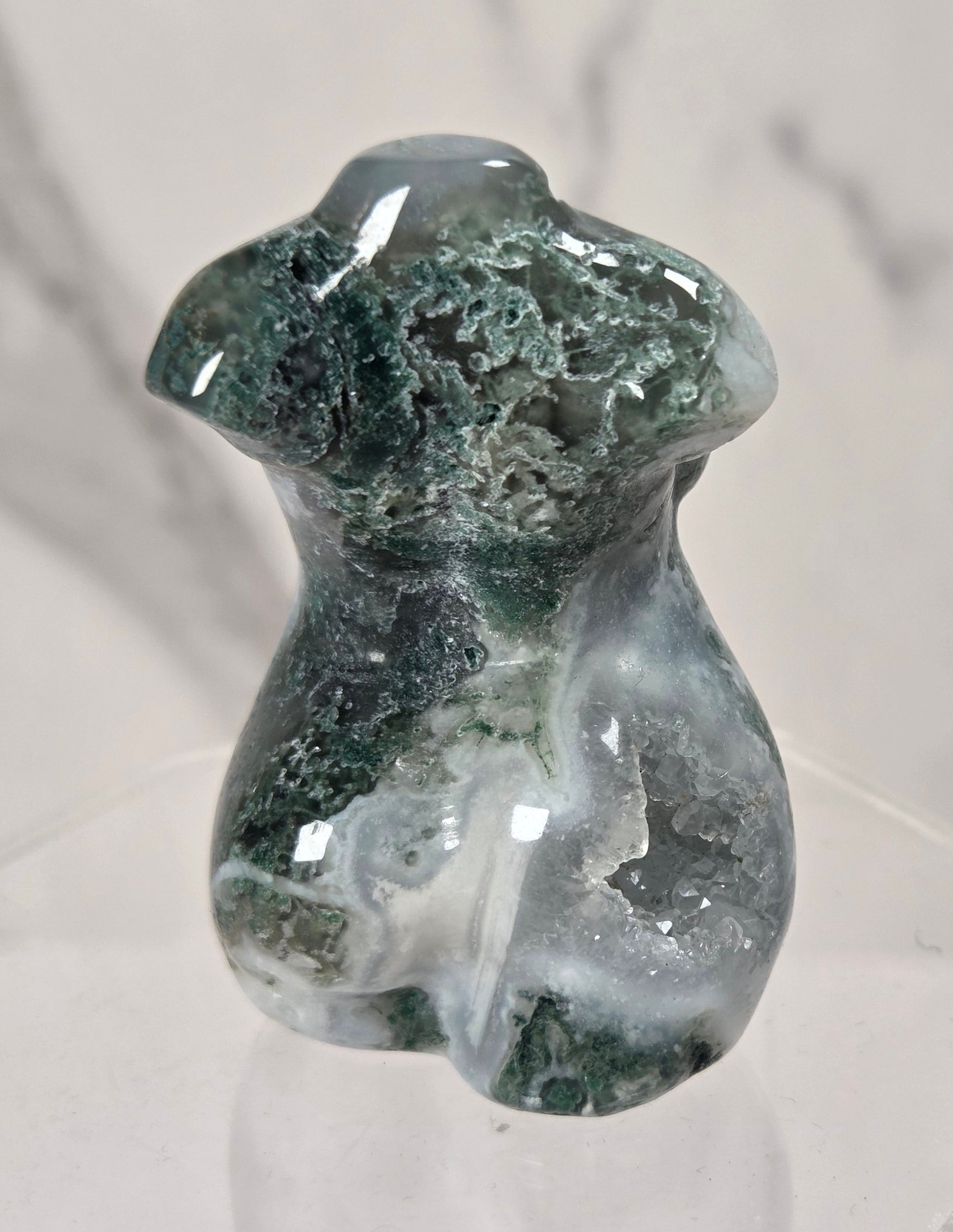 A small polished stone figurine shaped like a female torso, featuring dark green and white marbled patterning, displayed on a clear acrylic stand against a light marble-patterned background.