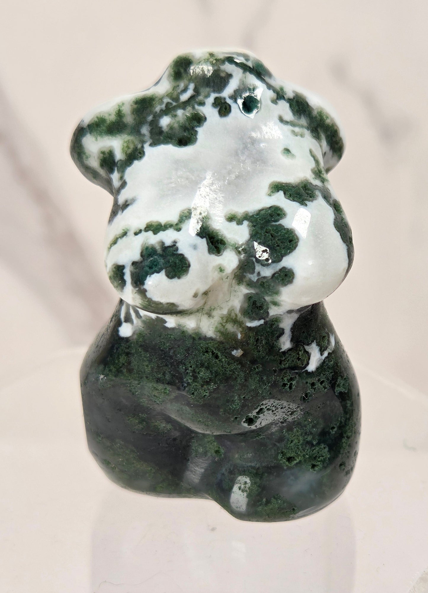 A small polished stone figurine shaped like a female torso, featuring dark green and white marbled patterning, displayed on a clear acrylic stand against a light marble-patterned background.