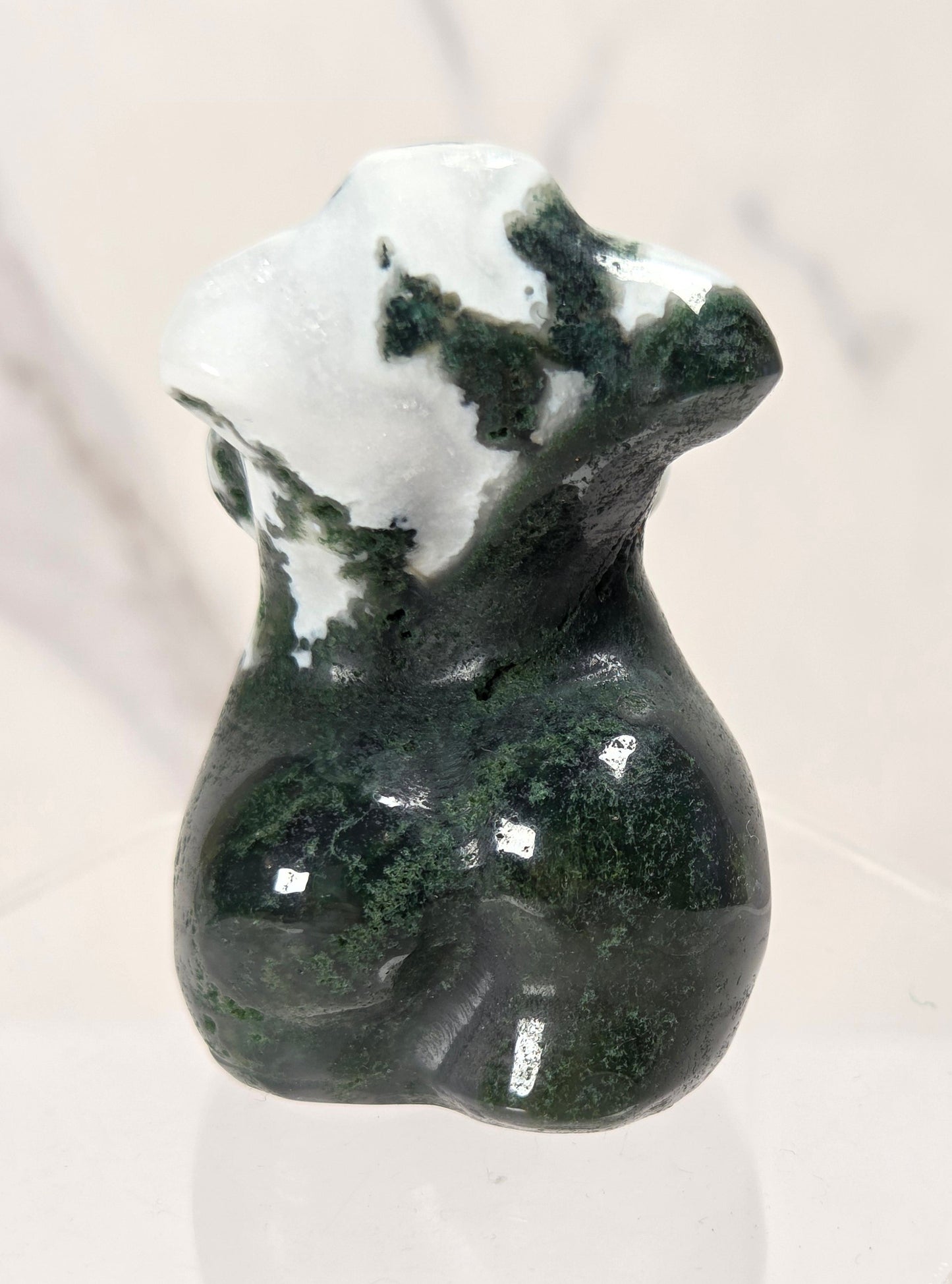 A small polished stone figurine shaped like a female torso, featuring dark green and white marbled patterning, displayed on a clear acrylic stand against a light marble-patterned background.