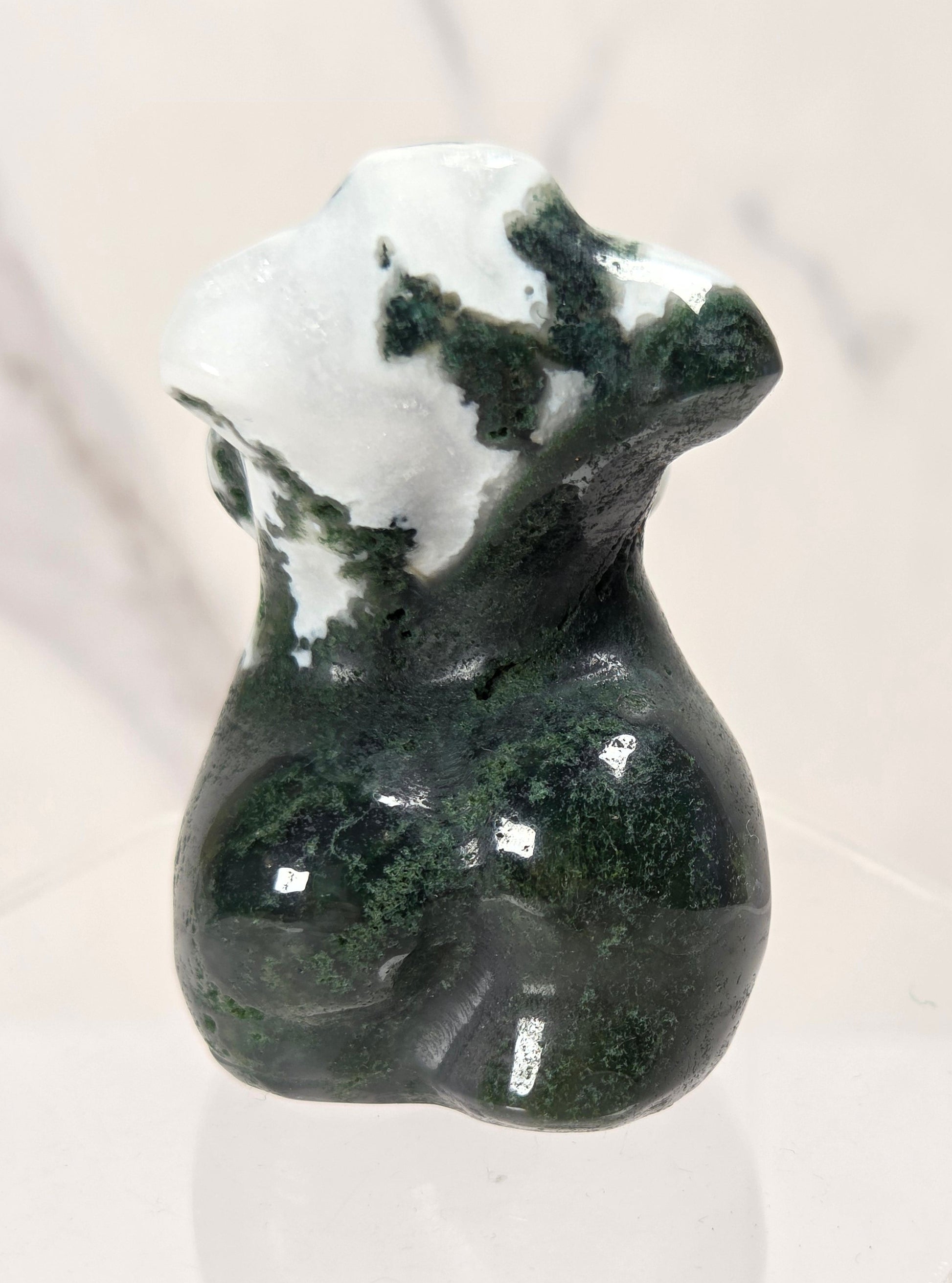 A small polished stone figurine shaped like a female torso, featuring dark green and white marbled patterning, displayed on a clear acrylic stand against a light marble-patterned background.