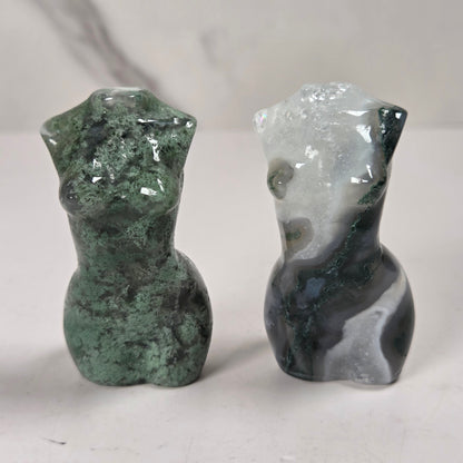 Two small polished stone figurines shaped like stylized female torso busts, one green marbled and one white and dark gray marbled, standing on a light surface against a light gray marble-patterned background.
