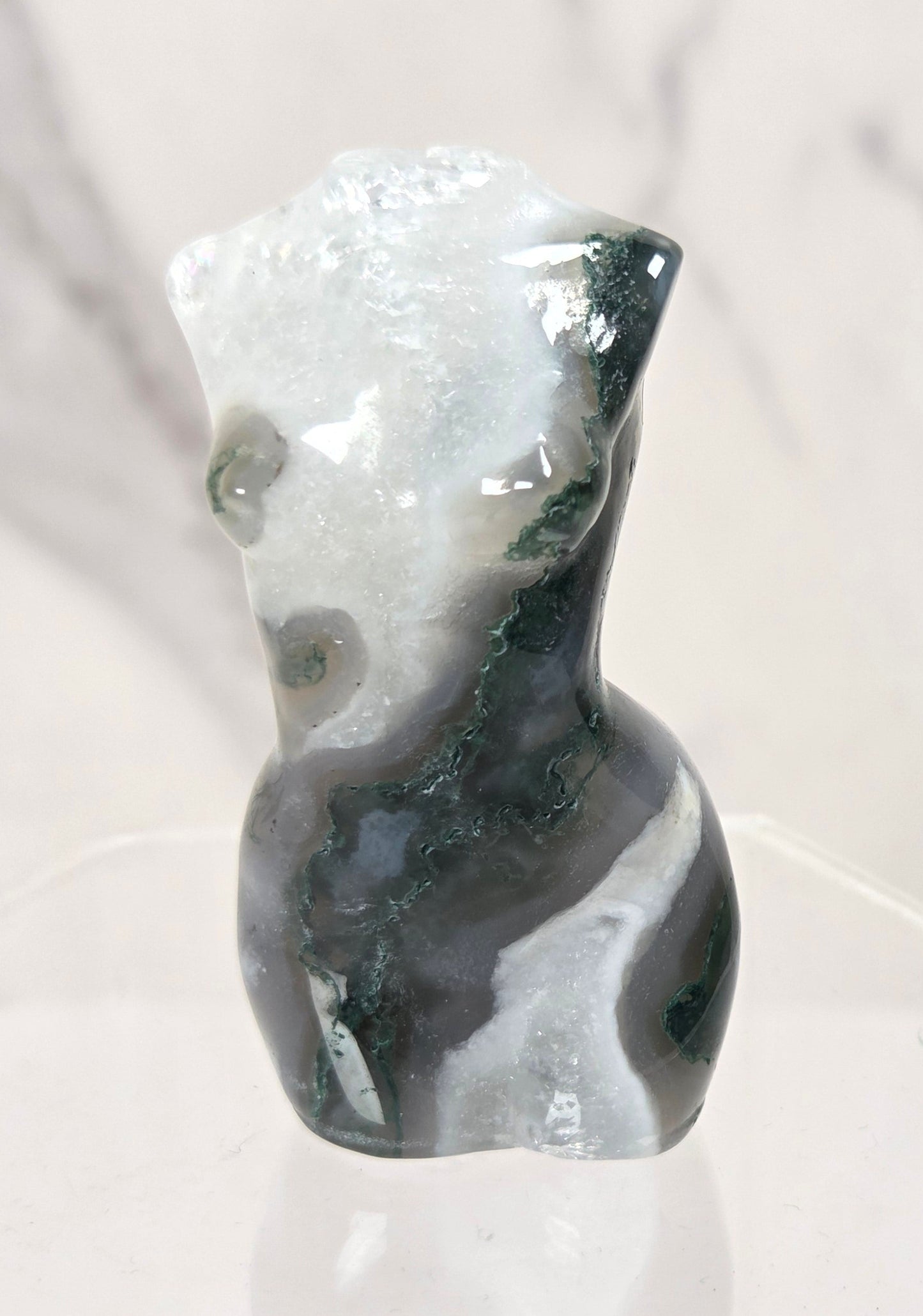 A small abstract torso-shaped stone sculpture with marbled black, white, and gray tones, displayed on a clear hexagonal pedestal against a light marble-look background.