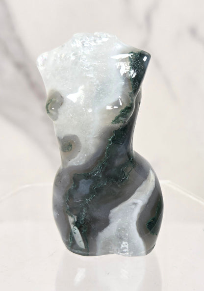 A small abstract torso-shaped stone sculpture with marbled black, white, and gray tones, displayed on a clear hexagonal pedestal against a light marble-look background.