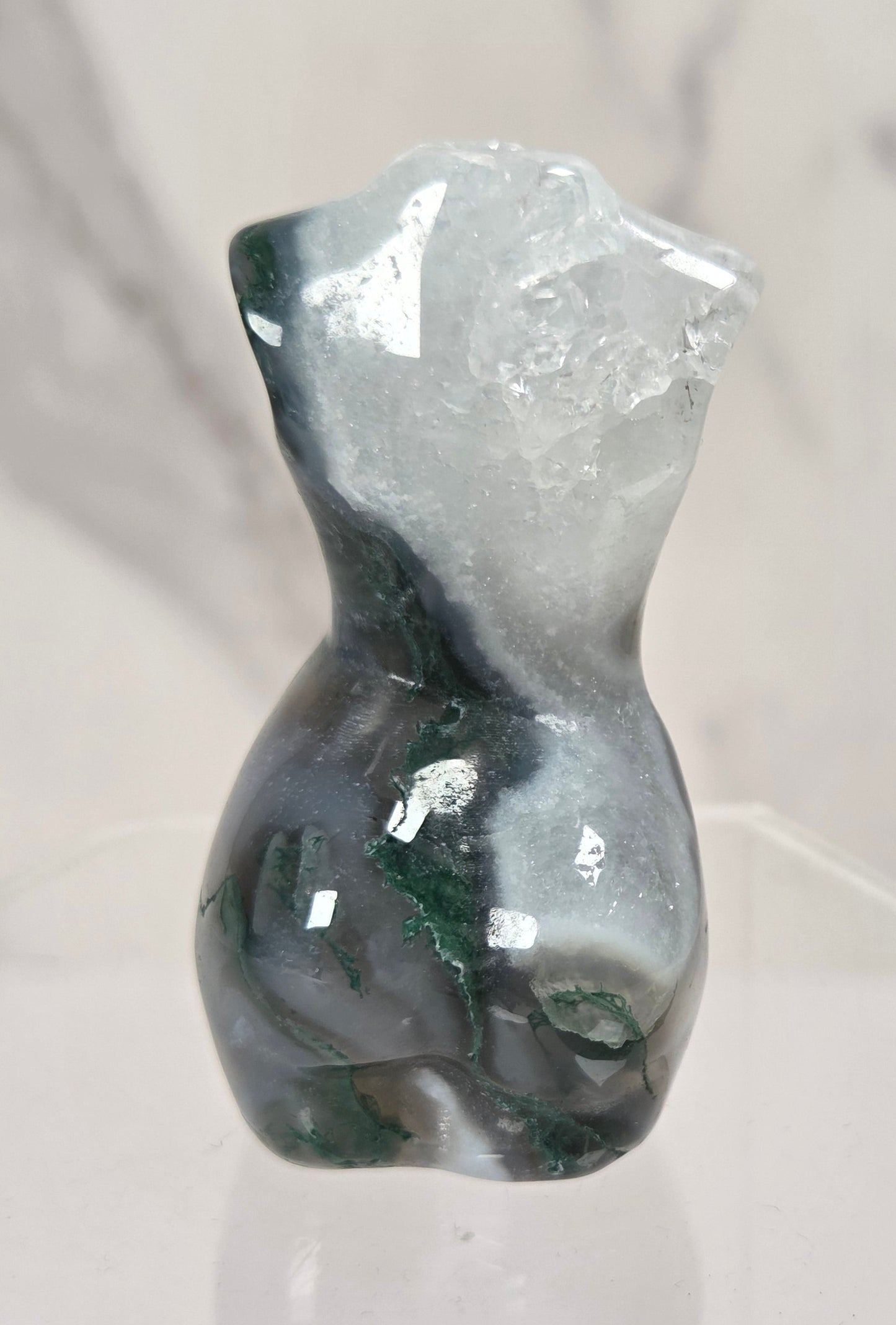 A small polished stone figurine shaped like a female torso, featuring dark green and white marbled patterning, displayed on a clear acrylic stand against a light marble-patterned background.