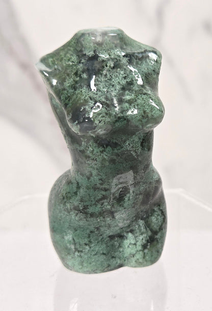 A small green stone figurine with a mottled, textured surface, displayed on a clear acrylic stand against a light gray marble-patterned background.
