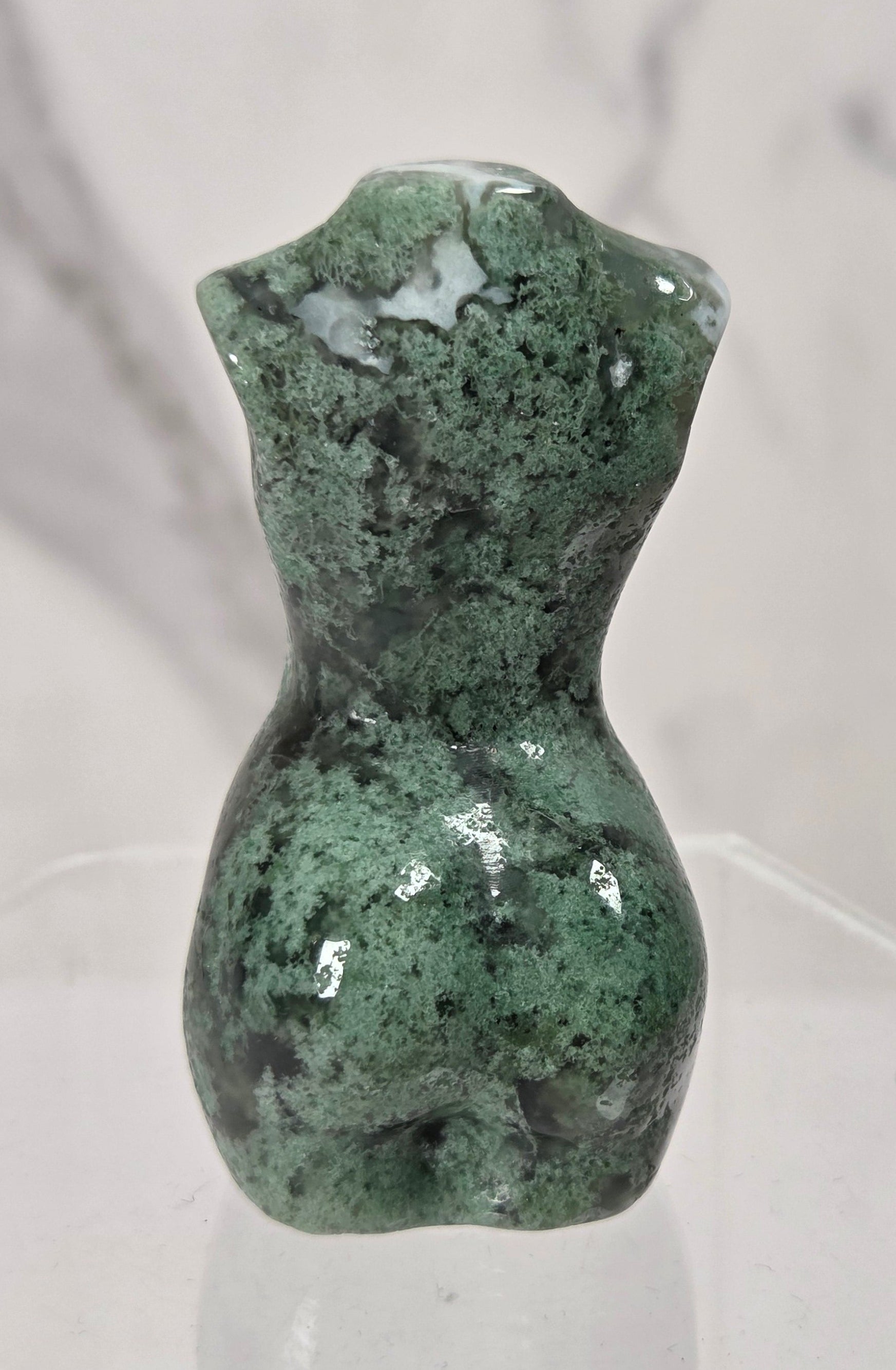 A small sculpted green stone torso figurine with a glossy finish, displayed on a clear acrylic hexagonal stand against a light marble-patterned background.
