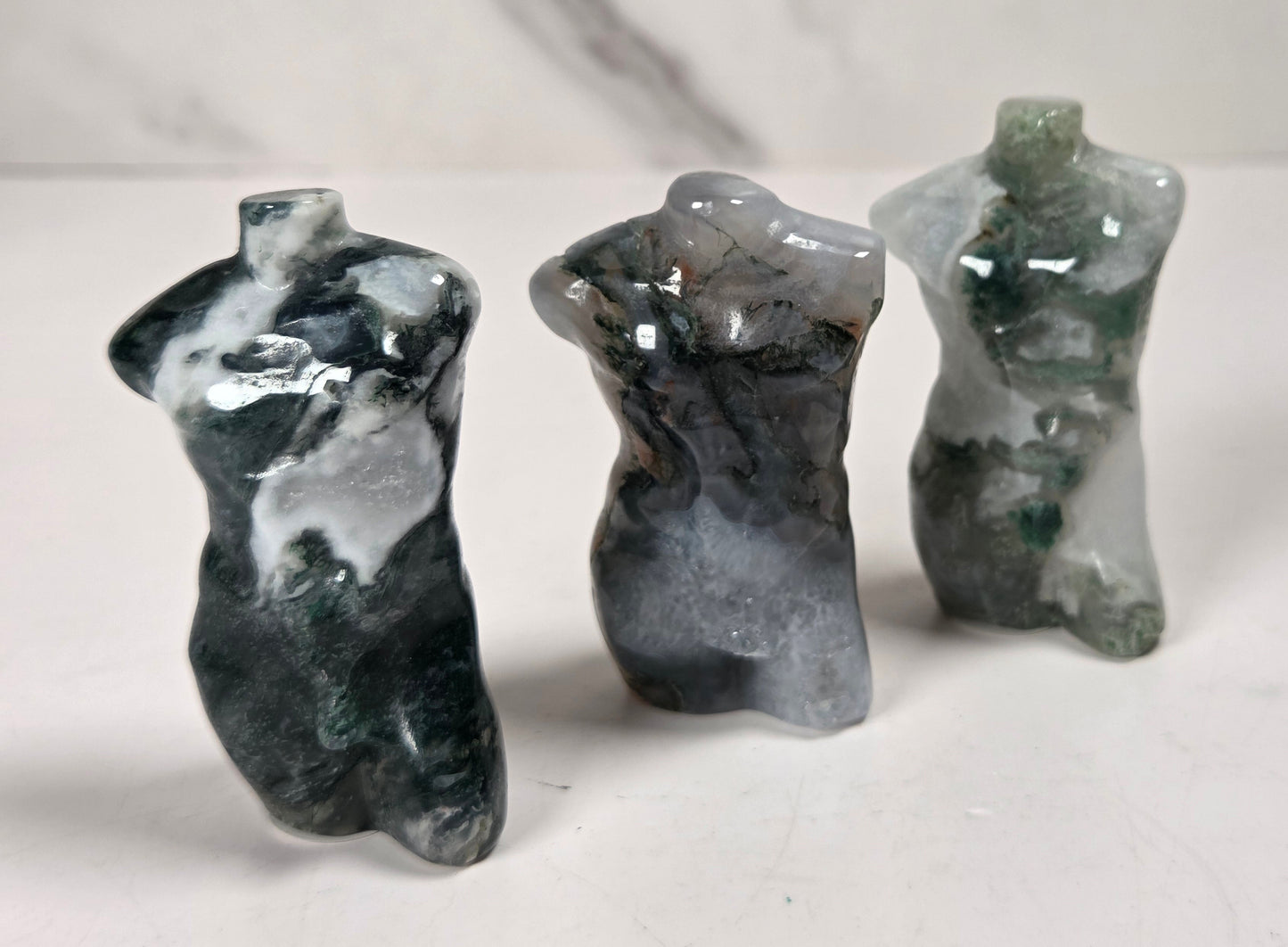 Three small abstract figurines shaped like stylized female torsos, made from polished stone or resin, standing on a white surface against a light gray marble-look background.