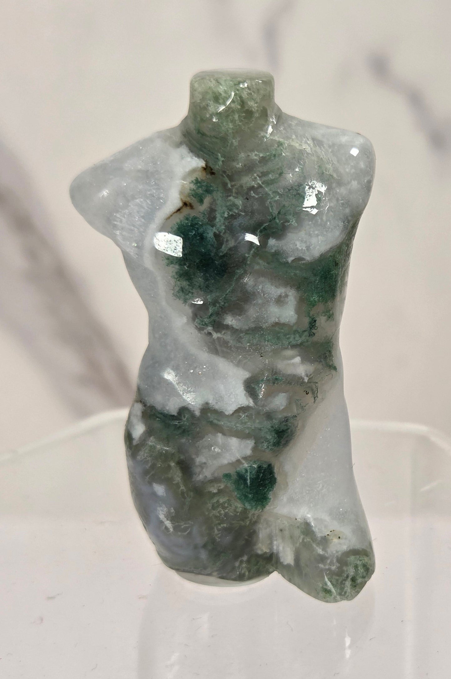 A small carved crystal or gemstone torso sculpture with a glossy finish, featuring natural green and milky white marbling, displayed on a clear acrylic stand against a light marble-patterned background.