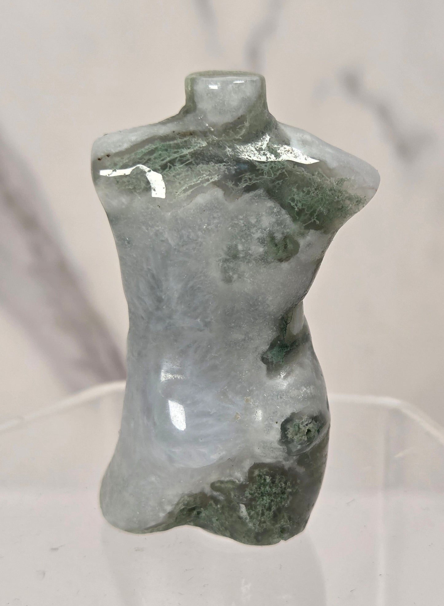 A small polished stone torso-shaped object with a glossy finish, featuring natural green and gray marbling, displayed on a clear acrylic stand against a light marble-patterned background.