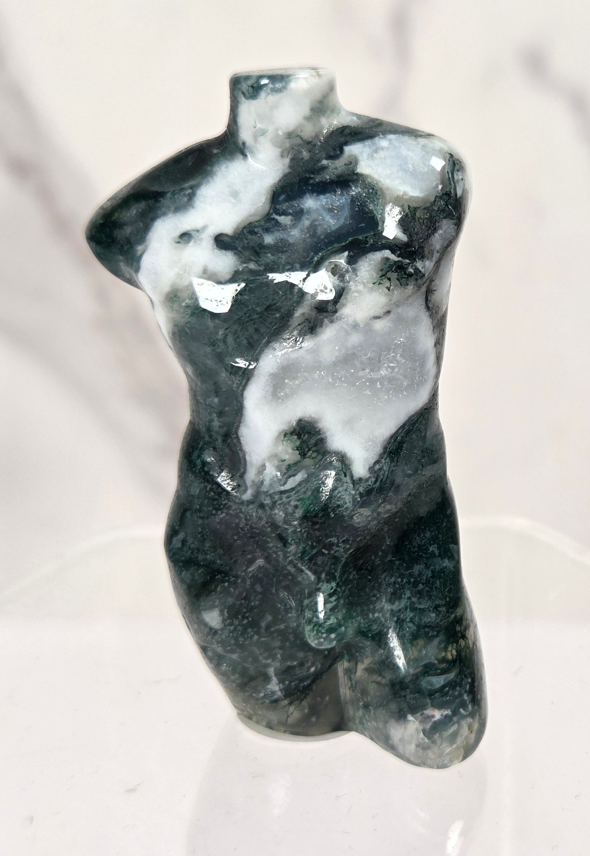 A small sculptural torso figurine made of polished dark green and white stone, displayed on a clear acrylic stand against a light marble-patterned background.