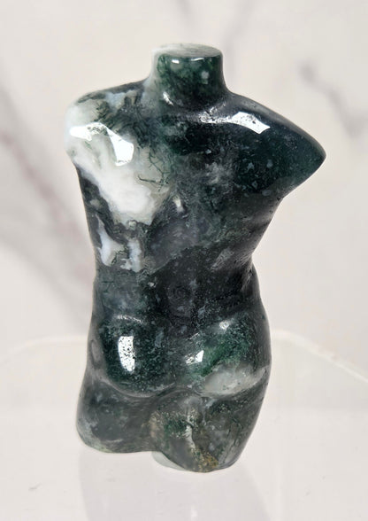 A small glossy torso-shaped sculpture made of dark green and white marbled stone, displayed on a clear acrylic stand against a light marble-like background.