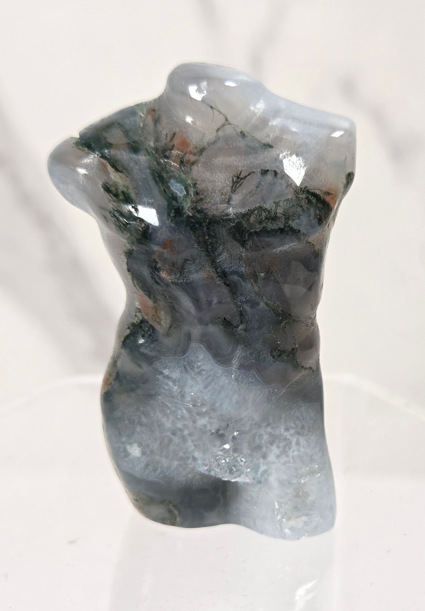 A small polished stone sculpture shaped like a torso, with a mottled mix of gray, blue-gray, black, and subtle green and brown mineral patterns, displayed on a clear acrylic stand against a light marble-patterned background.