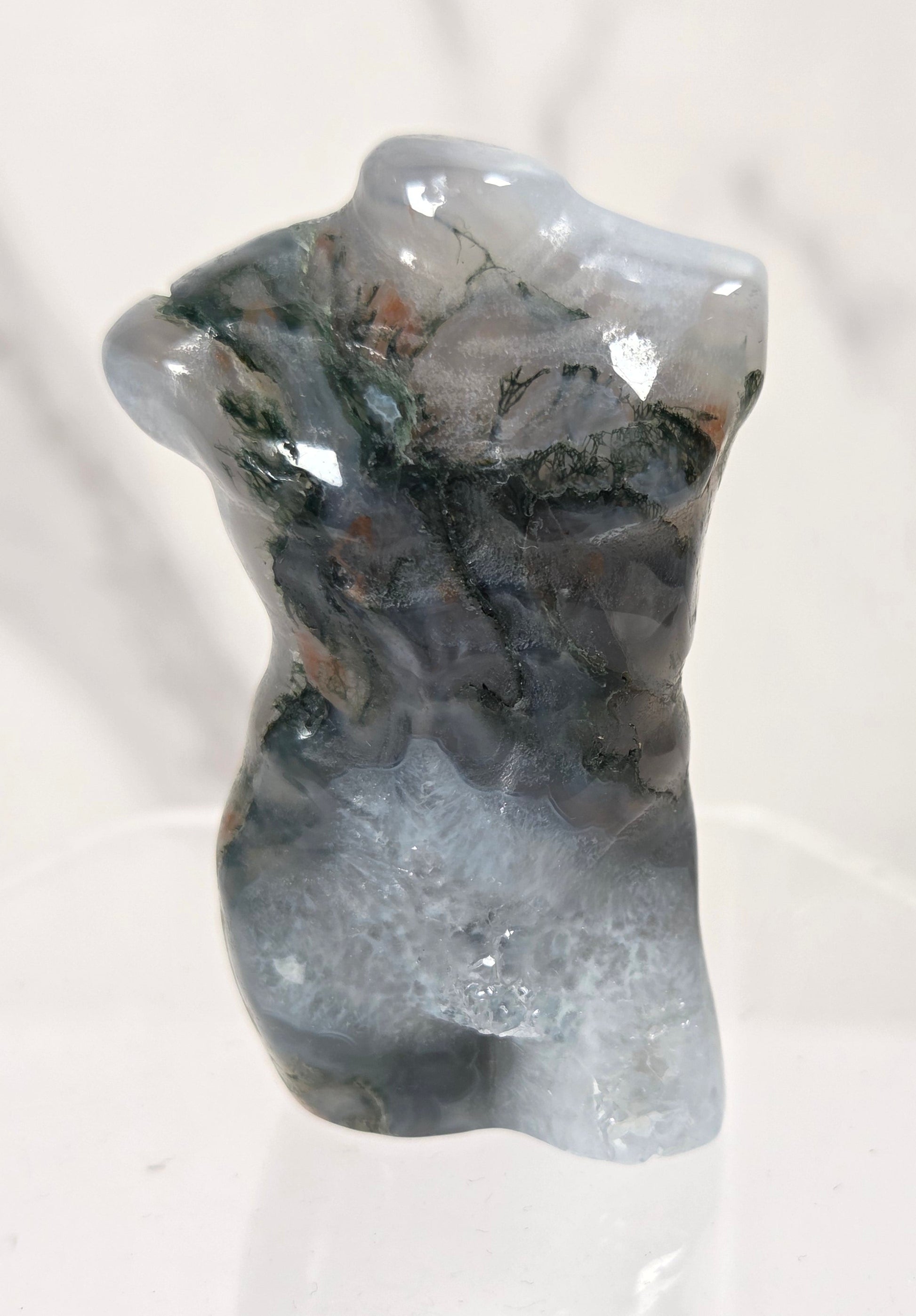 A small polished stone sculpture shaped like a torso, with a mottled mix of gray, blue-gray, black, and subtle green and brown mineral patterns, displayed on a clear acrylic stand against a light marble-patterned background.