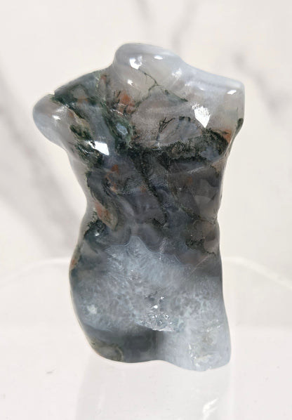 A small polished stone sculpture shaped like a torso, with a mottled mix of gray, blue-gray, black, and subtle green and brown mineral patterns, displayed on a clear acrylic stand against a light marble-patterned background.