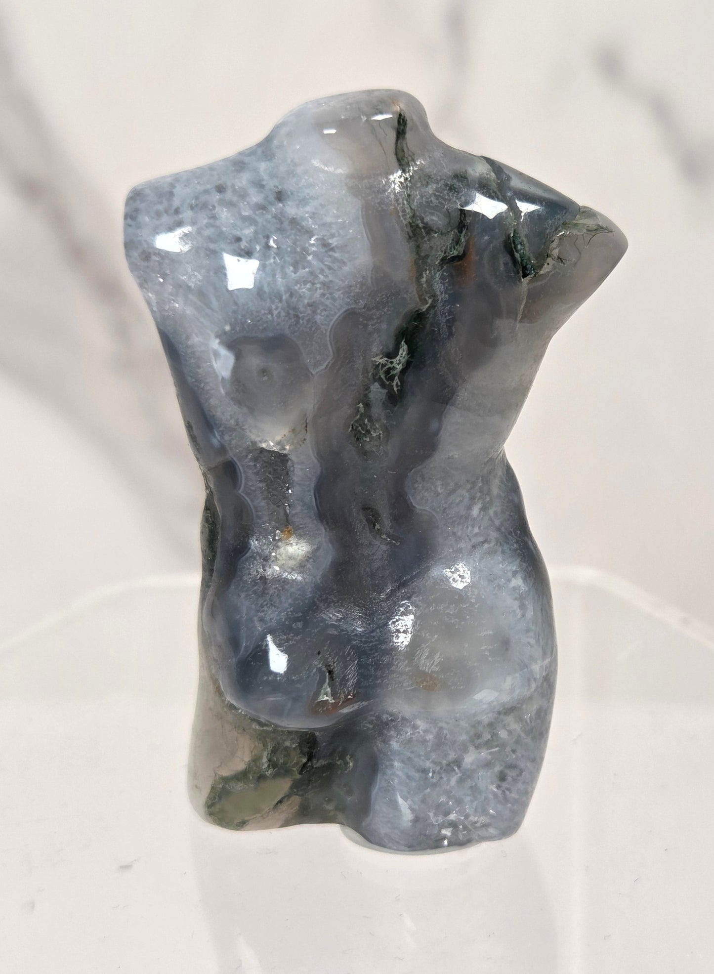 A small polished gray stone torso sculpture displayed on a clear acrylic stand against a light marble-patterned background.