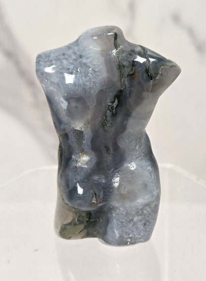 A small polished gray stone torso sculpture displayed on a clear acrylic stand against a light marble-patterned background.