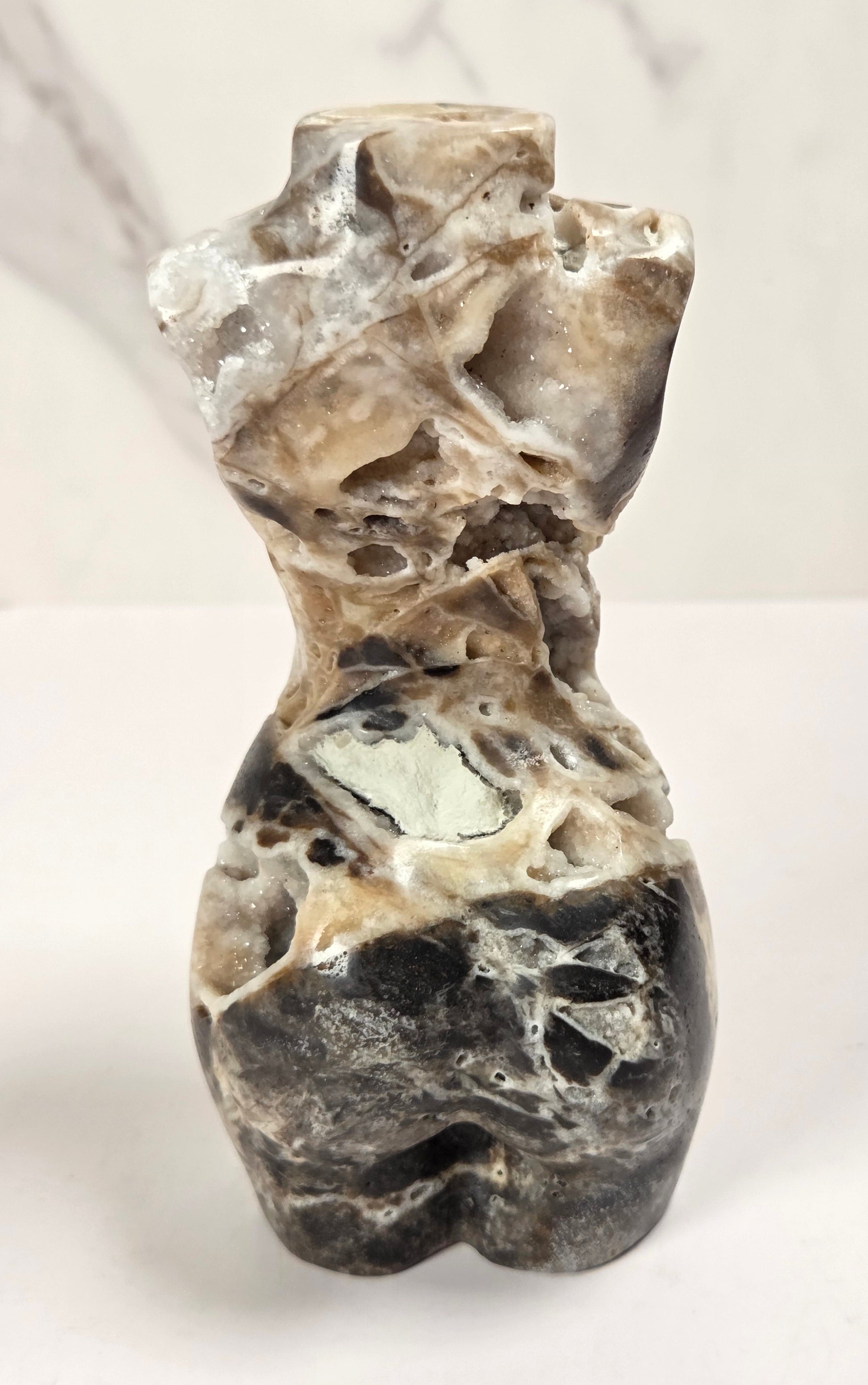 A small abstract stone sculpture shaped like a stylized torso, made from polished marbled stone with black, gray, beige, and white veining, photographed on a white surface against a light gray marble-look background.