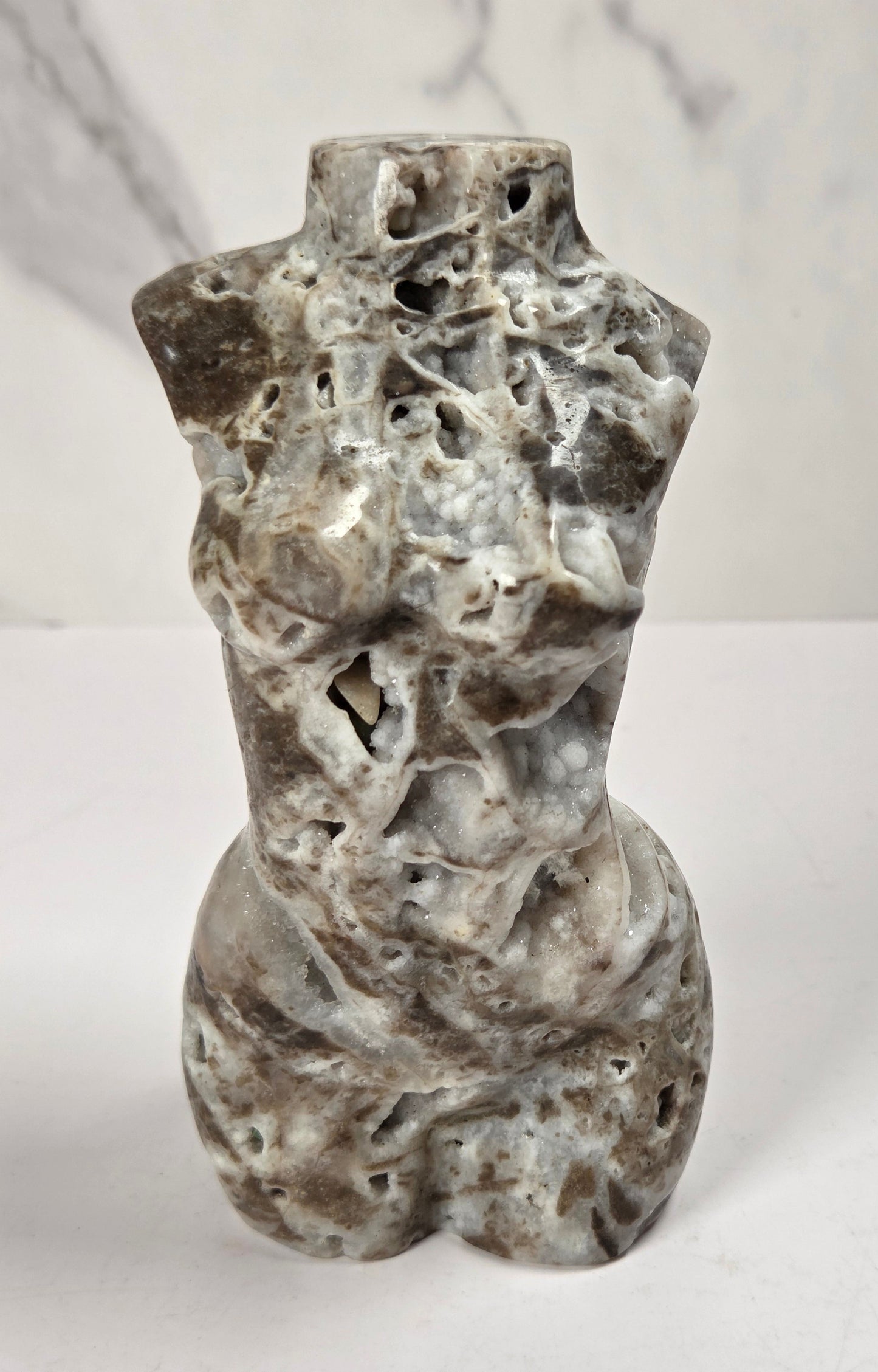 A small sculptural torso made from natural porous stone, featuring a mottled gray, white, and brown marbled pattern, standing on a white surface against a light gray marble-patterned background.