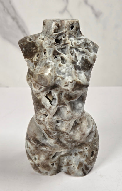 A small sculptural torso made from natural porous stone, featuring a mottled gray, white, and brown marbled pattern, standing on a white surface against a light gray marble-patterned background.