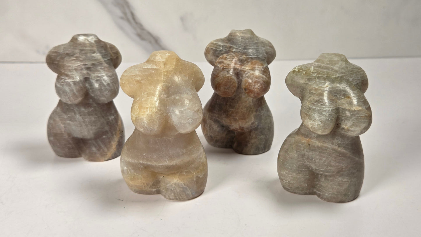 Four small carved stone figurines shaped like stylized human figures, shown standing on a white surface against a light gray marble-patterned background.