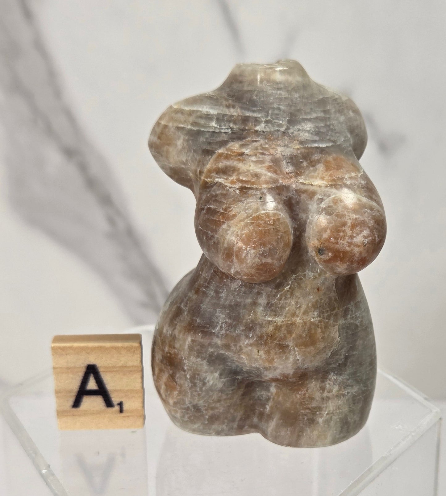 A small carved stone figurine with a weathered brown and gray texture, displayed on a clear acrylic stand next to a wooden Scrabble tile showing the letter A1, against a white marble-patterned background.