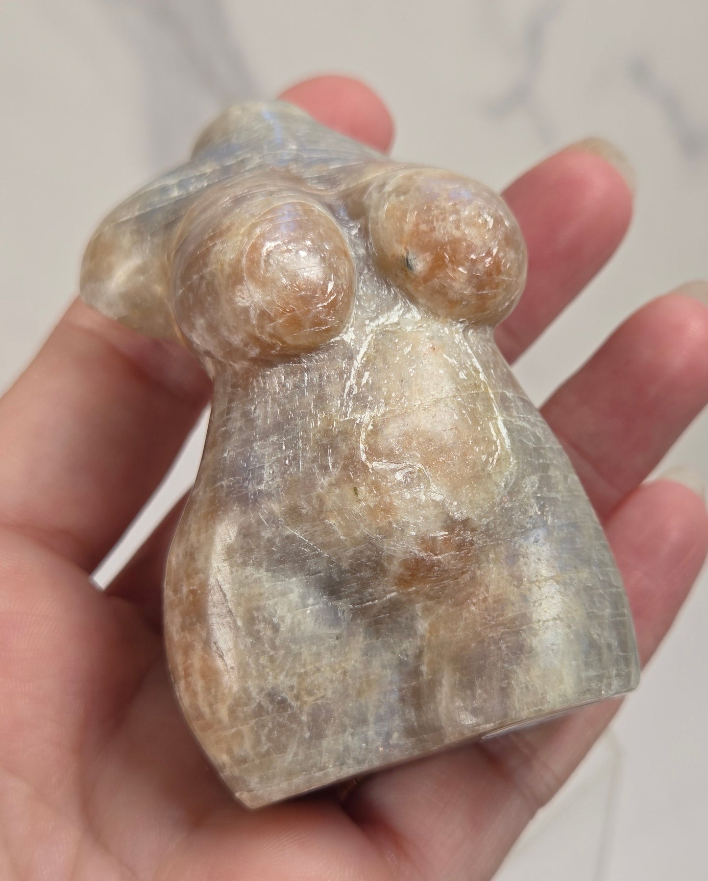 A hand holding a small carved figurine resembling a stylized female form, made from a polished stone with natural beige, brown, and gray marbling.