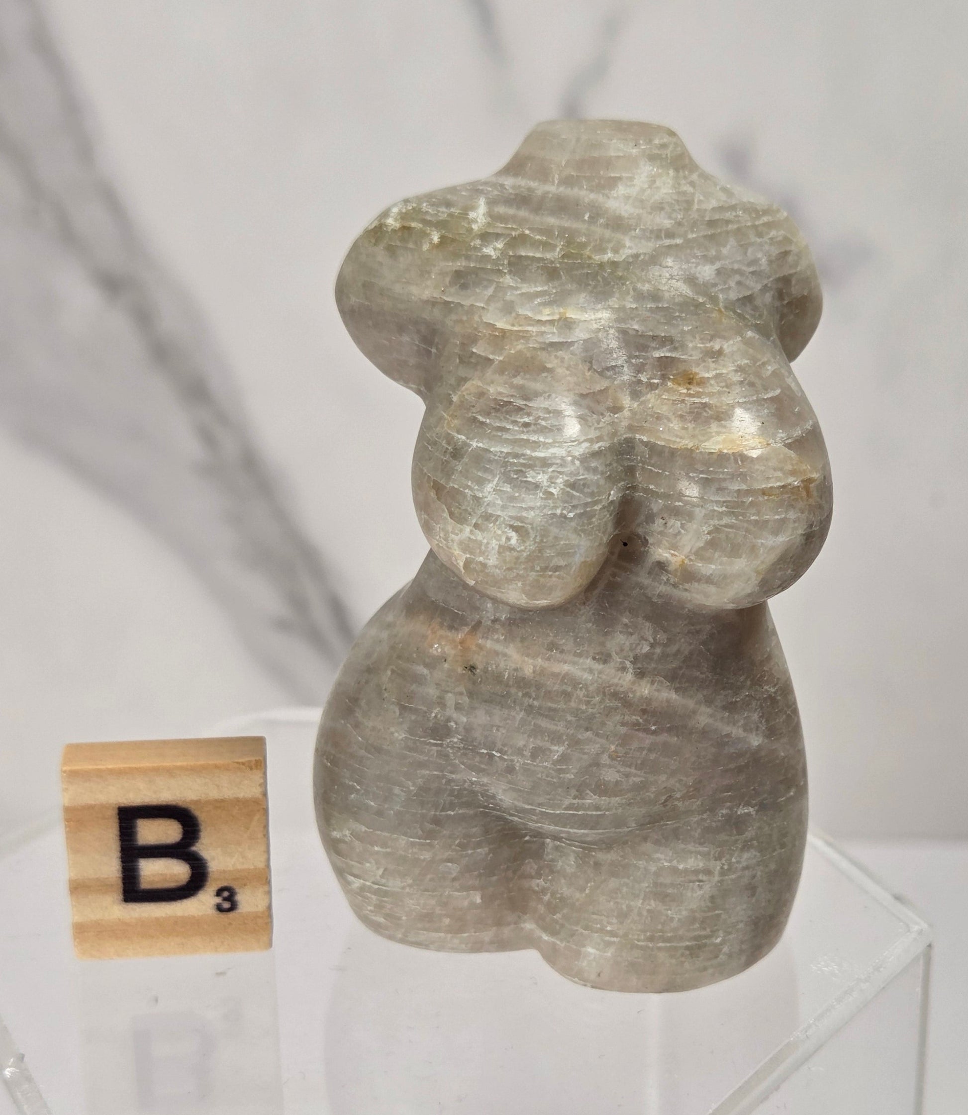 A small carved stone figurine with a stylized female form, shown on a clear acrylic pedestal next to a wooden Scrabble tile with the letter B.