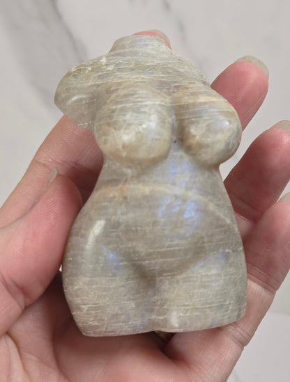 A hand holding a small carved stone figurine with a pearlescent, iridescent sheen. The figurine has a smooth, rounded organic shape resembling a stylized human torso.