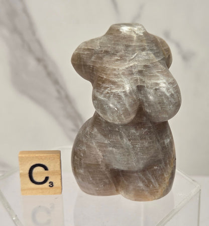 A small carved stone figurine with a smooth, rounded organic shape, displayed on a clear acrylic stand next to a wooden Scrabble tile showing the letter C. The background is a soft-focus light gray marble surface.