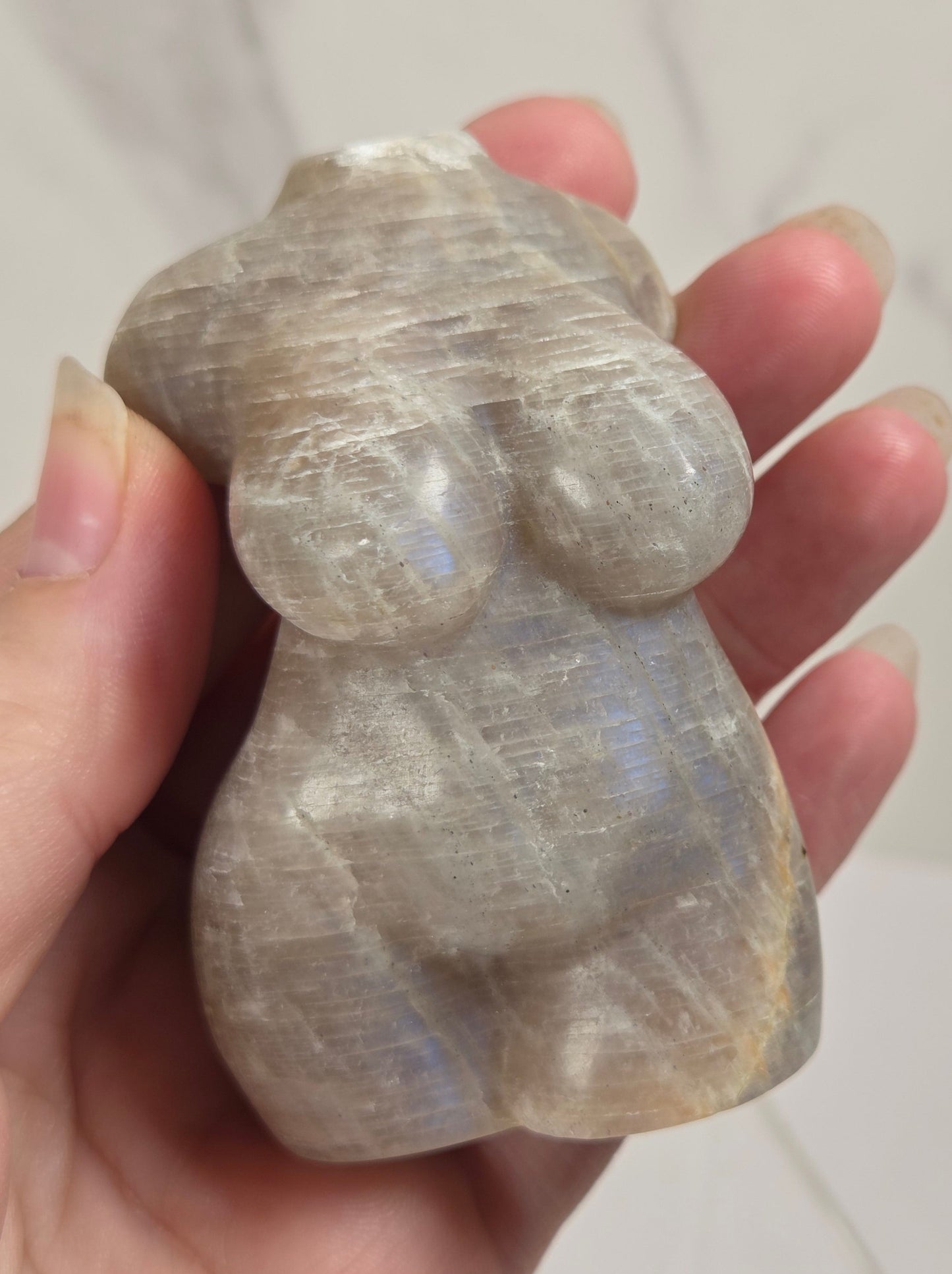 A hand holding a small, abstract carved stone sculpture with a pearlescent, iridescent sheen. The background is a soft-focus light gray marble surface.