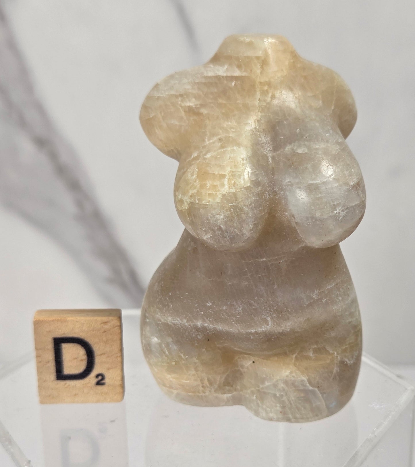 A small carved stone figurine with a smooth, polished surface, displayed on a clear acrylic stand next to a Scrabble tile showing the letter 'D'. The background is a soft-focus gray marble surface.