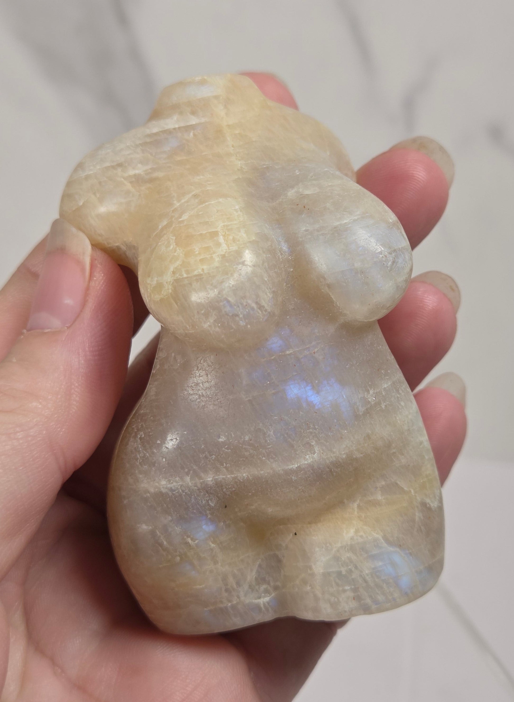 A hand holding a small carved stone figurine with a pearlescent, milky appearance and subtle blue iridescence, set against a light marble-patterned background.
