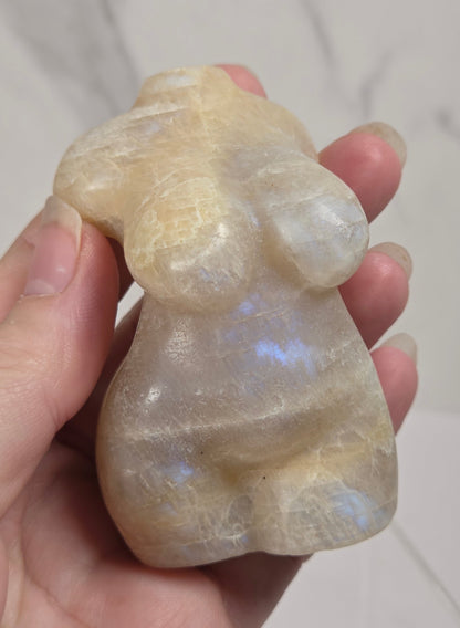A hand holding a small carved stone figurine with a pearlescent, milky appearance and subtle blue iridescence, set against a light marble-patterned background.