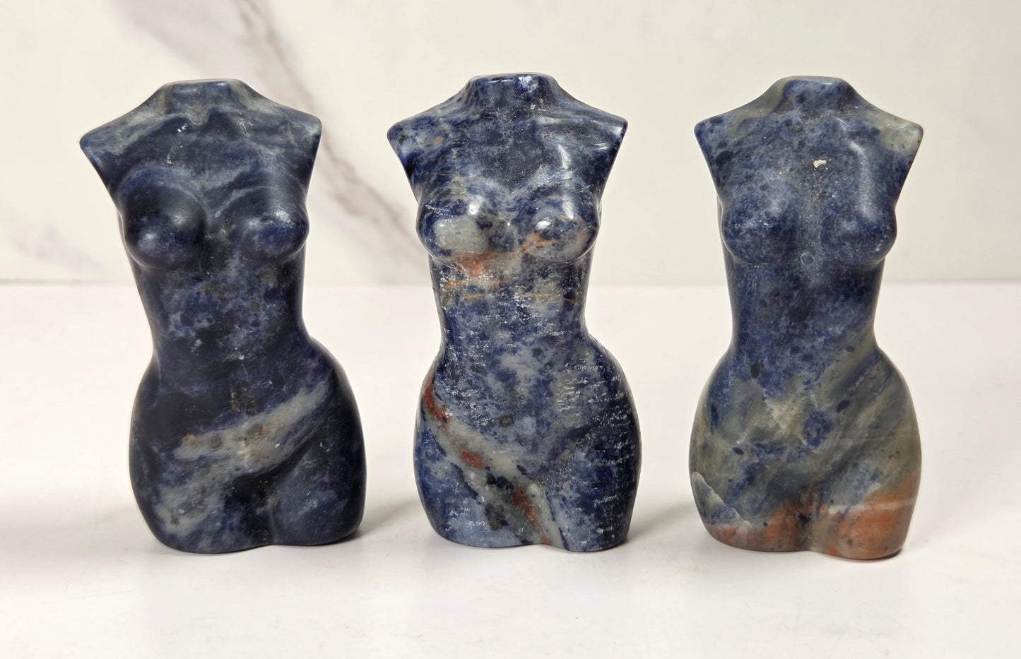 Three small carved stone figurines shaped like stylized female torso busts, shown standing side by side on a white surface against a light marble-look background.