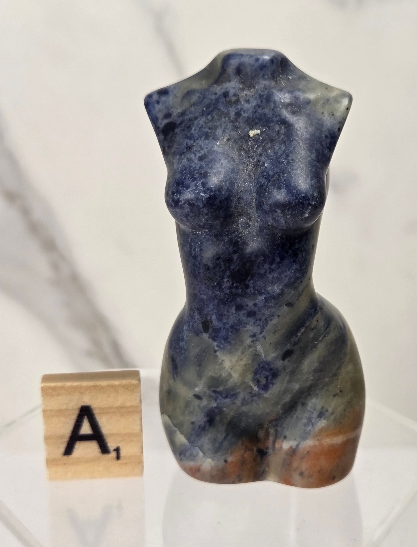 A small carved stone torso sculpture in deep blue lapis lazuli with natural marbling and brownish base tones, displayed on a clear acrylic stand next to a wooden Scrabble tile showing the letter A.