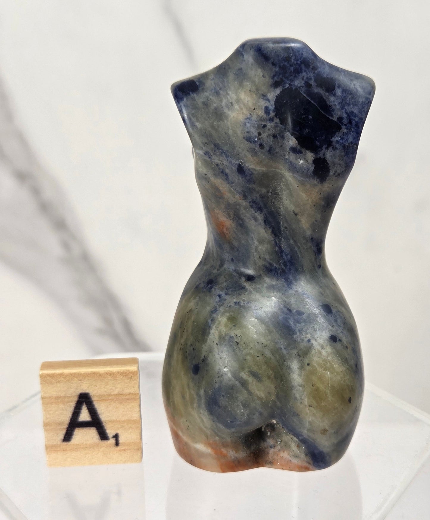 A small sculptural torso-shaped stone object in marbled blue, gray, olive, and brown tones, displayed on a clear acrylic stand next to a wooden letter tile showing 'A1', against a light marble-pattern background.