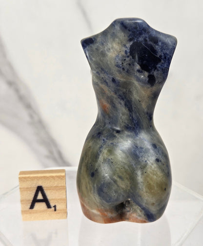 A small sculptural torso-shaped stone object in marbled blue, gray, olive, and brown tones, displayed on a clear acrylic stand next to a wooden letter tile showing 'A1', against a light marble-pattern background.