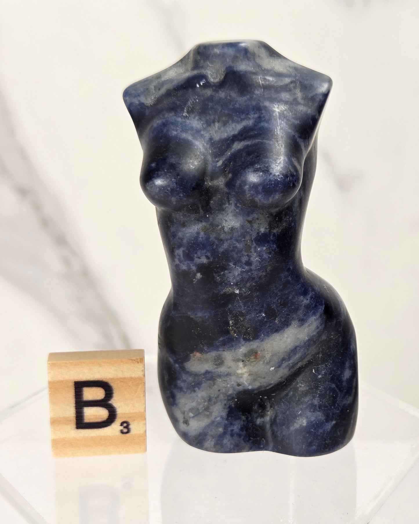 A small carved stone torso sculpture in deep blue-gray with natural marbling, displayed on a clear acrylic stand next to a wooden Scrabble tile showing the letter B.