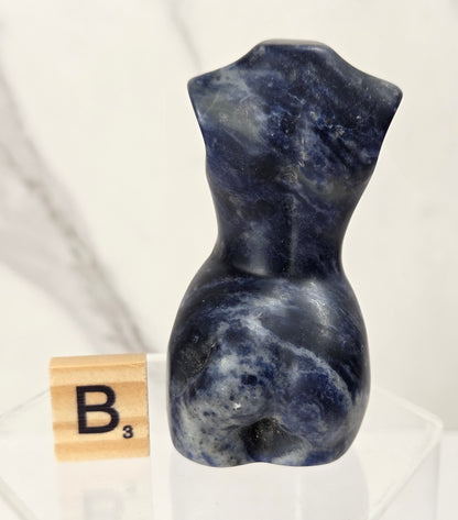 A small sculptural torso made of polished blue-gray stone with white marbling, displayed on a clear acrylic pedestal next to a wooden Scrabble tile showing the letter B.