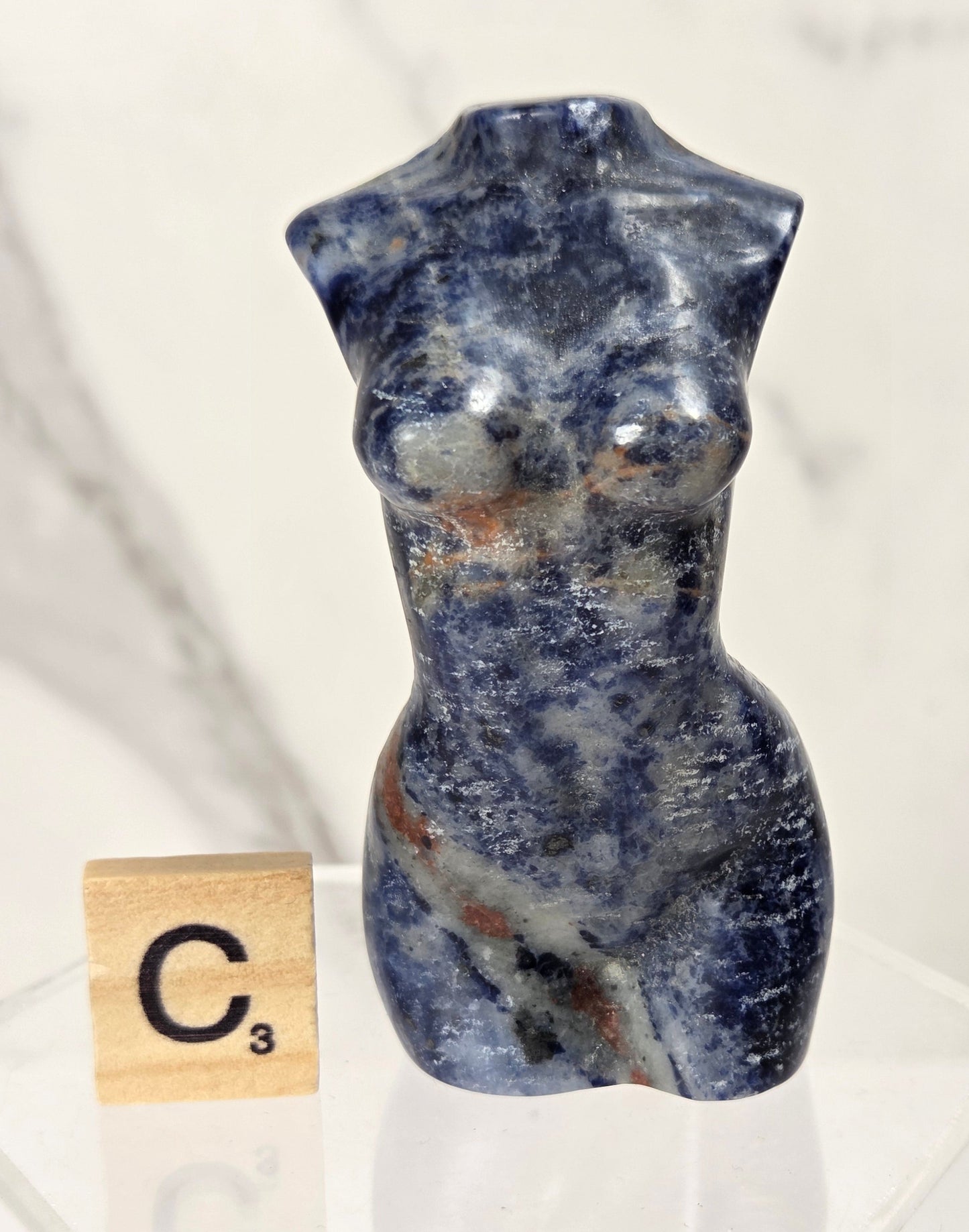 A small carved stone torso sculpture in deep blue with white and brown marbling, displayed on a clear acrylic stand next to a Scrabble tile showing C3.
