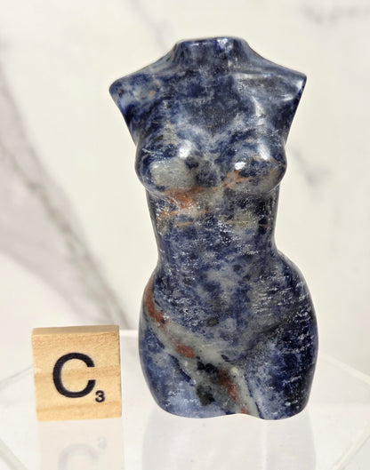 A small carved stone torso sculpture in deep blue with white and brown marbling, displayed on a clear acrylic stand next to a Scrabble tile showing C3.