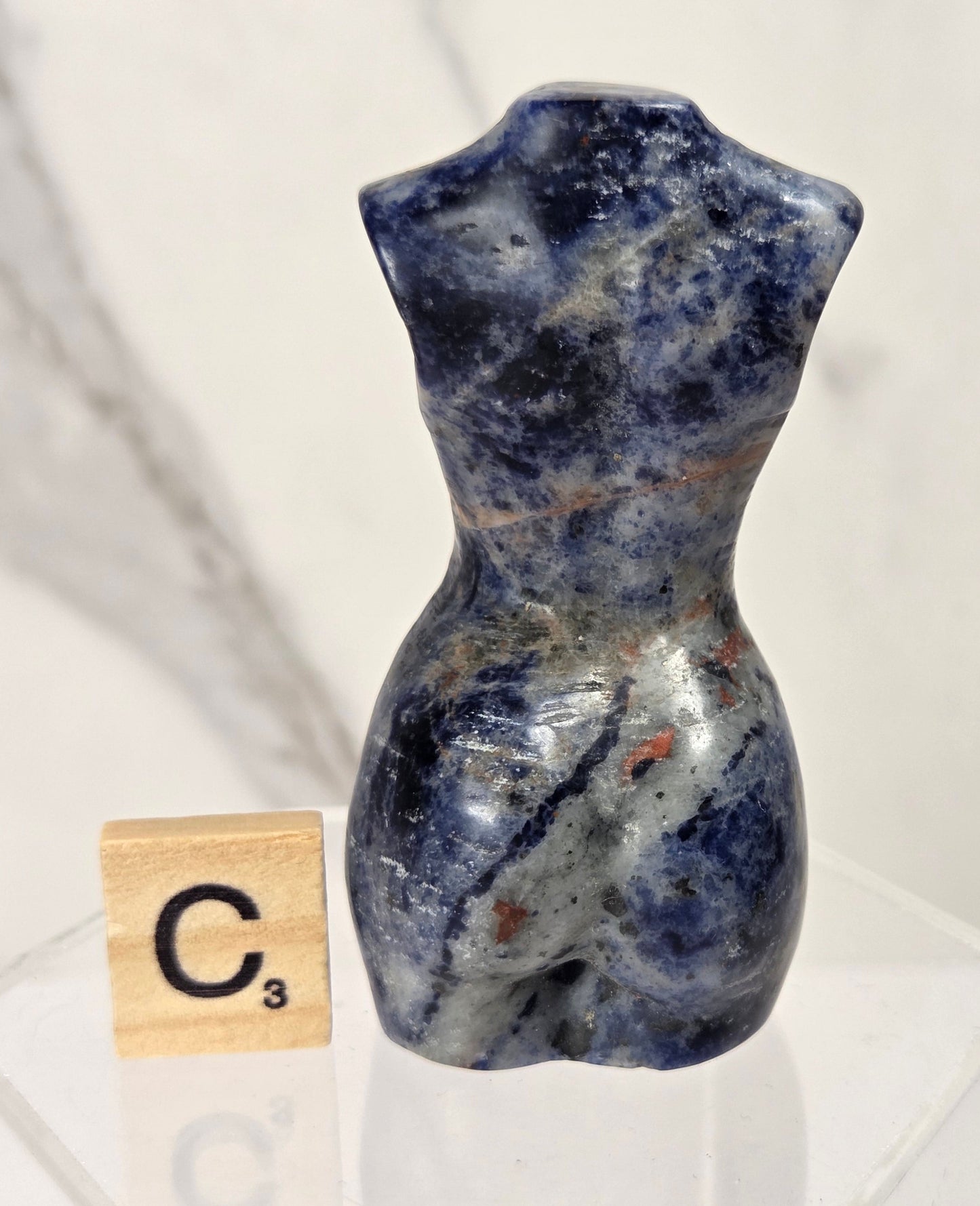 A small polished blue-and-white stone torso sculpture displayed on a clear acrylic stand next to a wooden Scrabble tile showing the letter C.