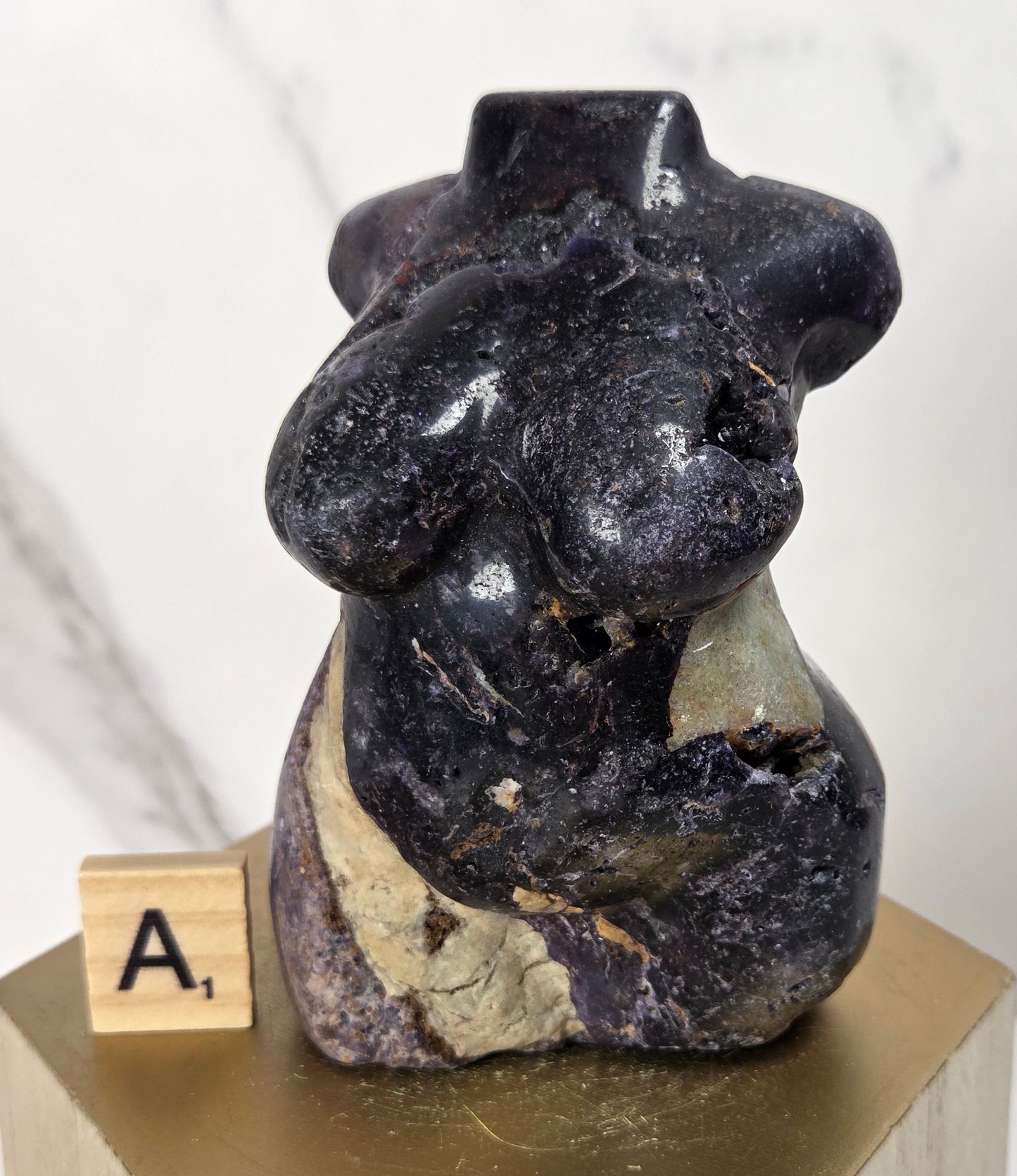 A small carved stone sculpture of a seated nude female figure, shown on a gold-colored pedestal with a wooden letter tile displaying 'A1'. The sculpture is made of dark polished black stone with natural beige and gray veining, set against a light marble background.