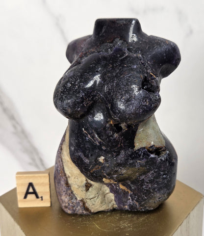 A small carved stone sculpture of a seated nude female figure, shown on a gold-colored pedestal with a wooden letter tile displaying 'A1'. The sculpture is made of dark polished black stone with natural beige and gray veining, set against a light marble background.