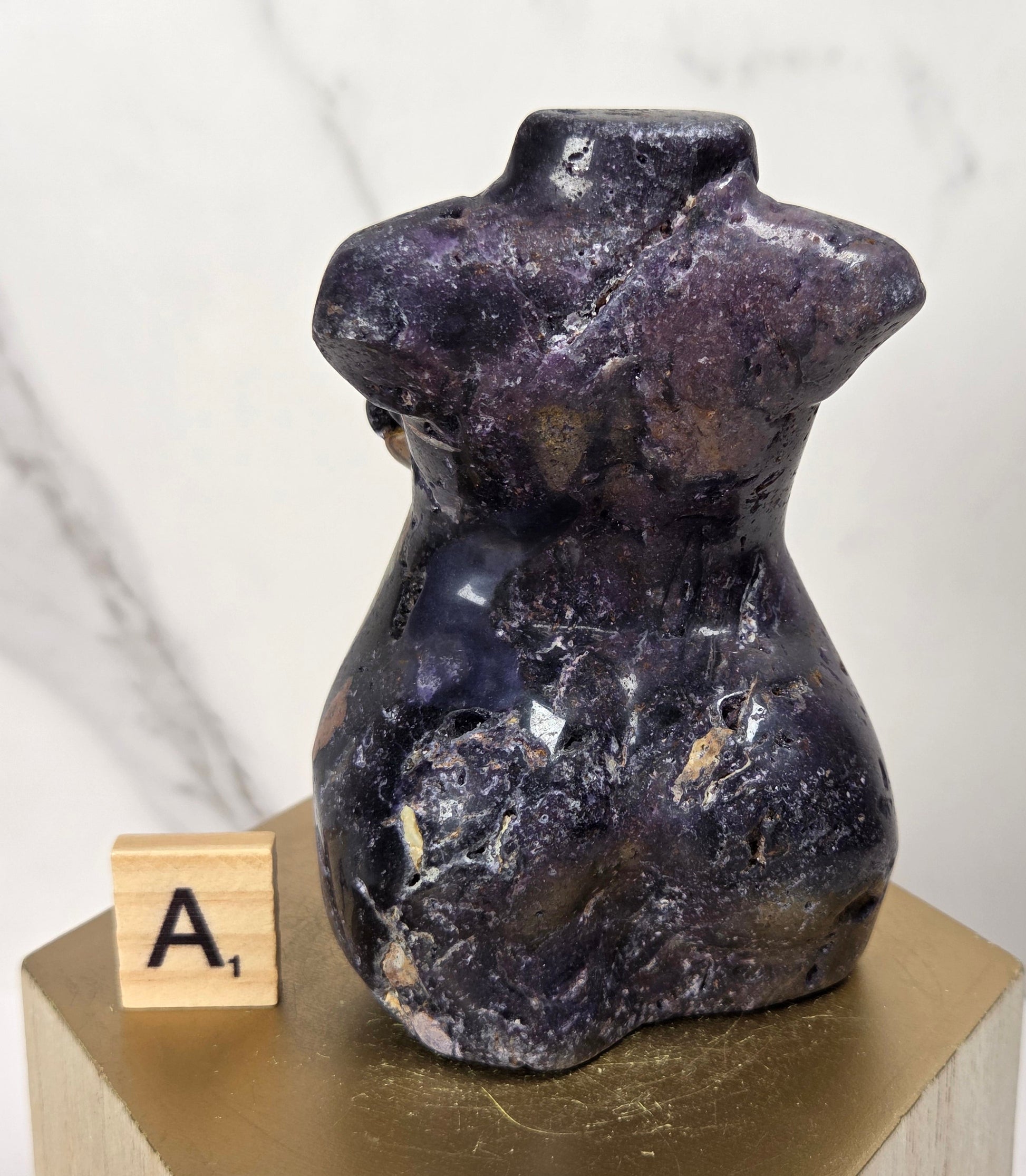 A small polished purple crystal torso sculpture displayed on a gold hexagonal pedestal with a wooden letter tile showing 'A1' beside it, against a light marble-patterned background.