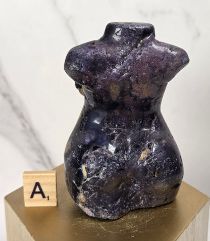 A small polished purple crystal torso sculpture displayed on a gold hexagonal pedestal with a wooden letter tile showing 'A1' beside it, against a light marble-patterned background.