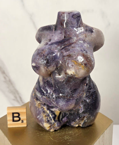 A polished purple amethyst figurine of a seated female form, displayed on a gold hexagonal pedestal with a wooden 'B' block tile next to it, against a light marble background.