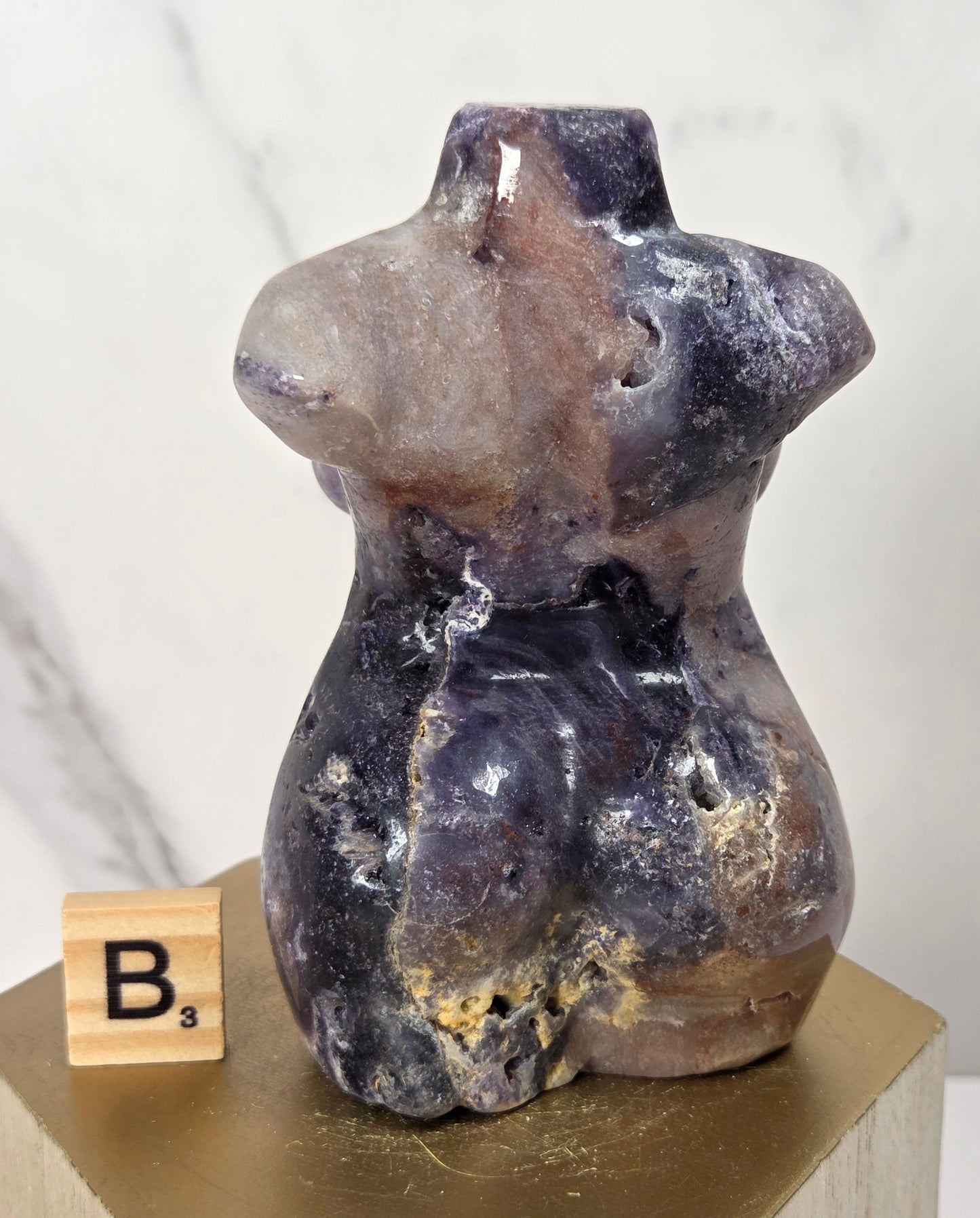 A small sculptural torso-shaped object made of polished stone in mottled purple, brown, and cream tones, displayed on a gold pedestal next to a wooden Scrabble tile.