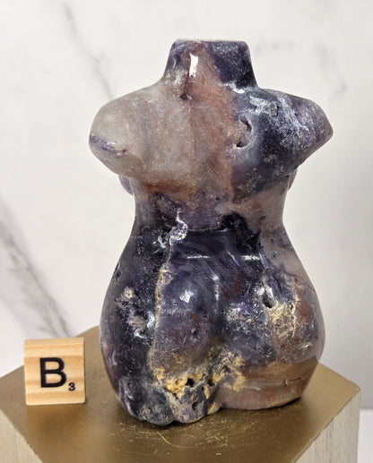 A small sculptural torso-shaped object made of polished stone in mottled purple, brown, and cream tones, displayed on a gold pedestal next to a wooden Scrabble tile.